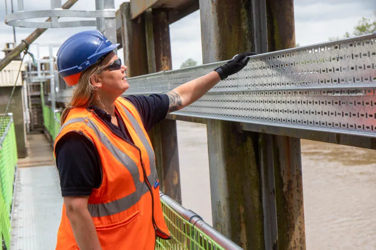 A woman in a hard hat and orange vest inspects a metal structure by a river, touching it with a gloved hand.