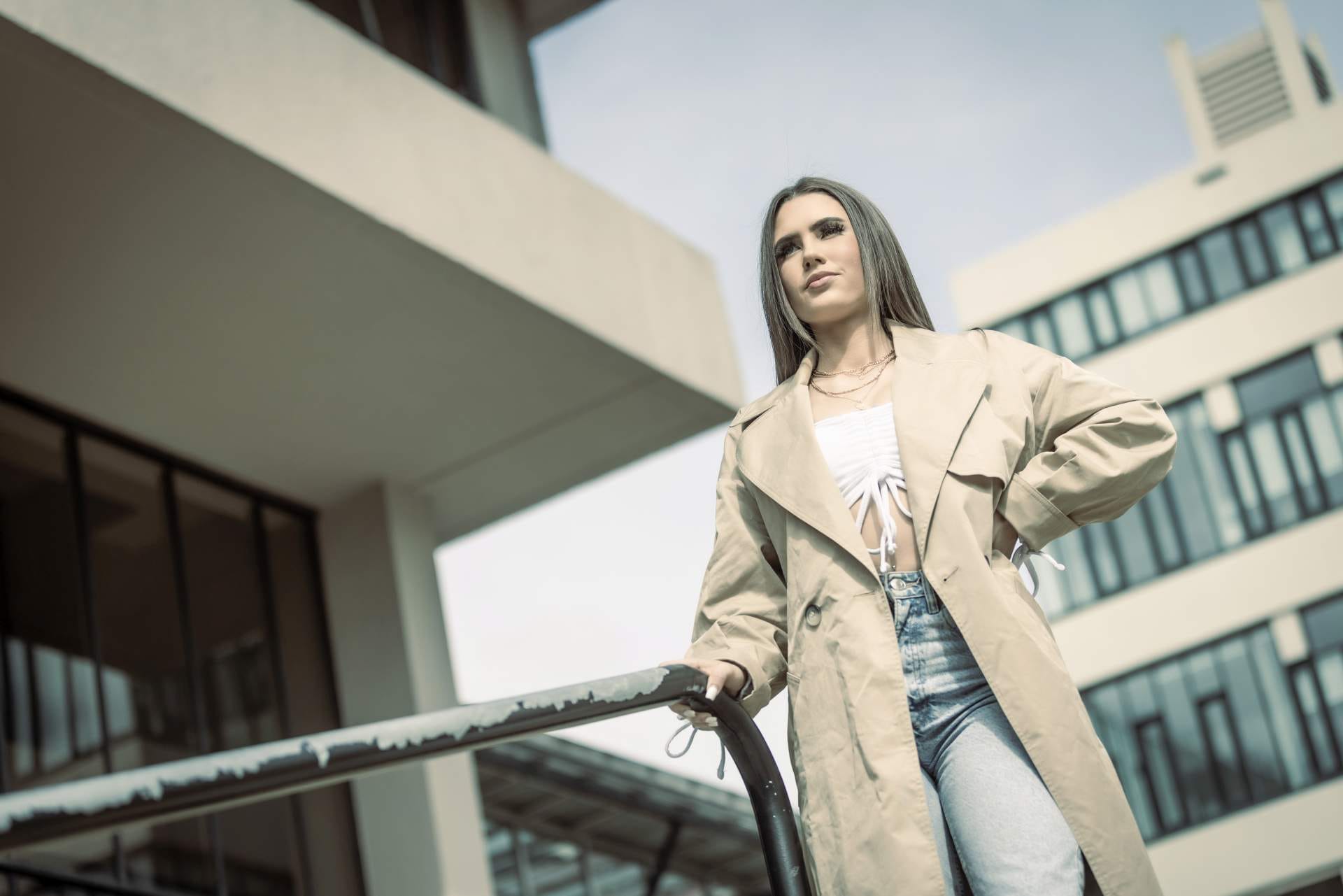 Woman in a beige trench coat and jeans stands confidently in front of modern buildings, hand resting on a metal rail.