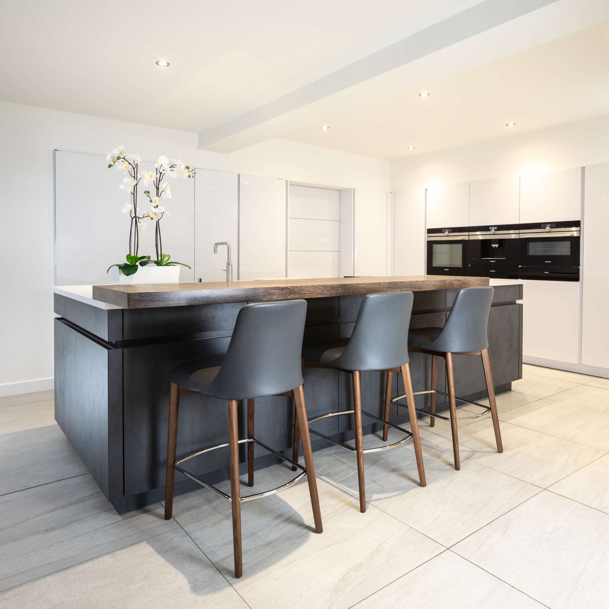 Modern kitchen with a large island featuring three barstools, white countertops, sleek cabinets, and a potted orchid.