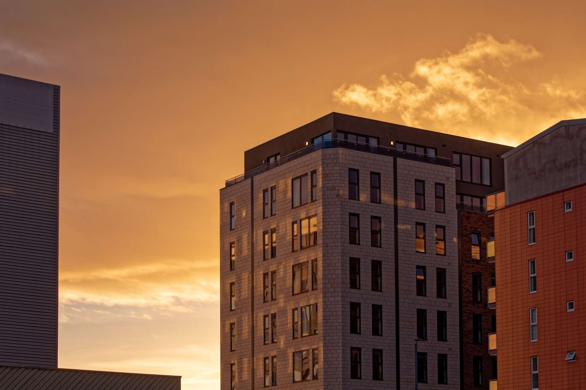 Modern apartment buildings against a vibrant orange sunset sky, with reflections in the windows and scattered clouds above.