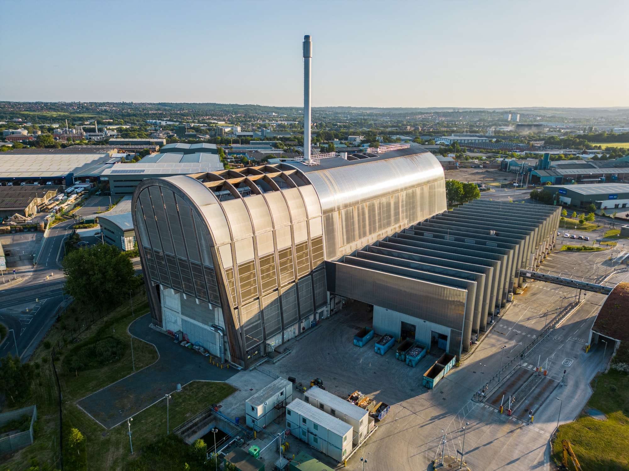 Aerial view of a large industrial waste-to-energy plant with a tall chimney, surrounded by other industrial buildings and roads.