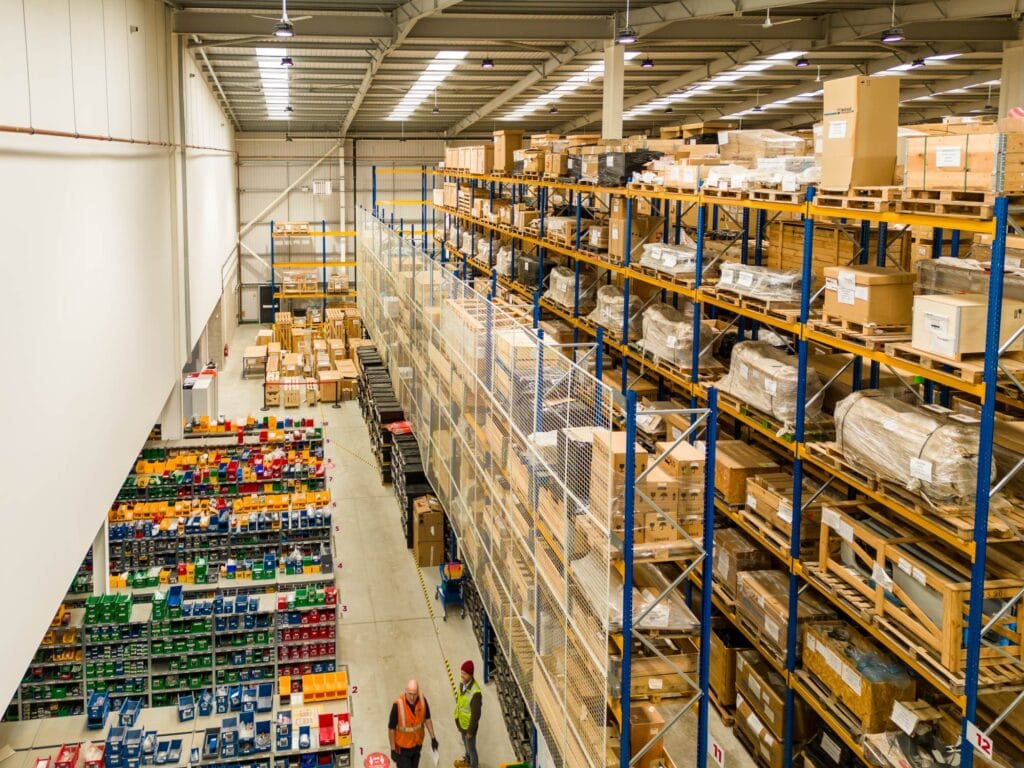 Large warehouse with tall shelves stacked with various boxes and parcels. Two workers in high-visibility vests at the bottom.