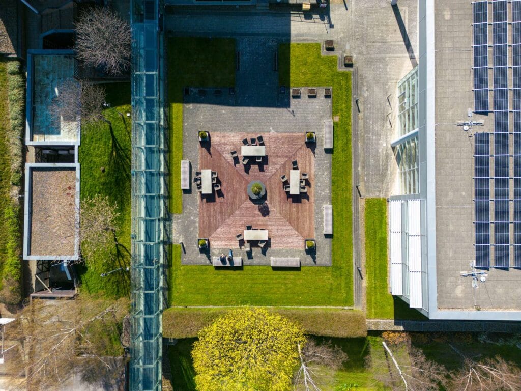 Aerial view of a rectangular courtyard with seating areas, surrounded by grass and modern buildings, including a solar-paneled roof.