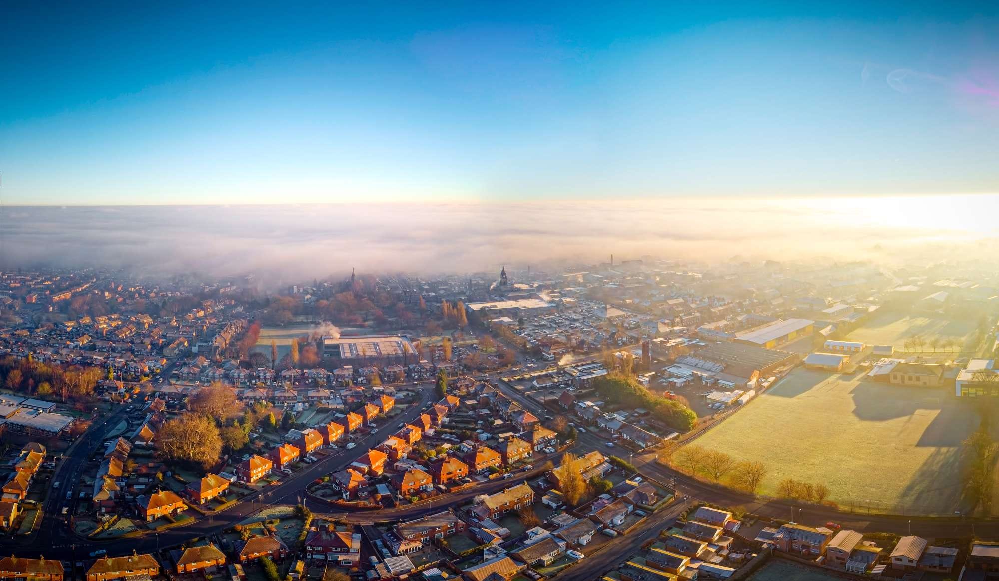 Aerial view of a town at sunrise with houses, greenery, and fog-covered buildings in the distance under a clear blue sky.