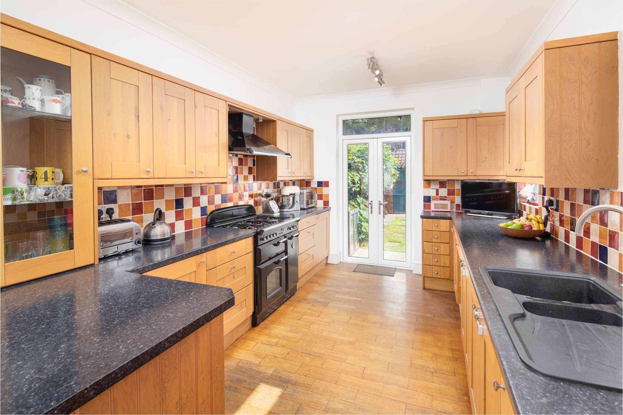 Modern kitchen with wooden cabinets, black countertops, colorful tiled backsplash, and a double door leading to a garden.