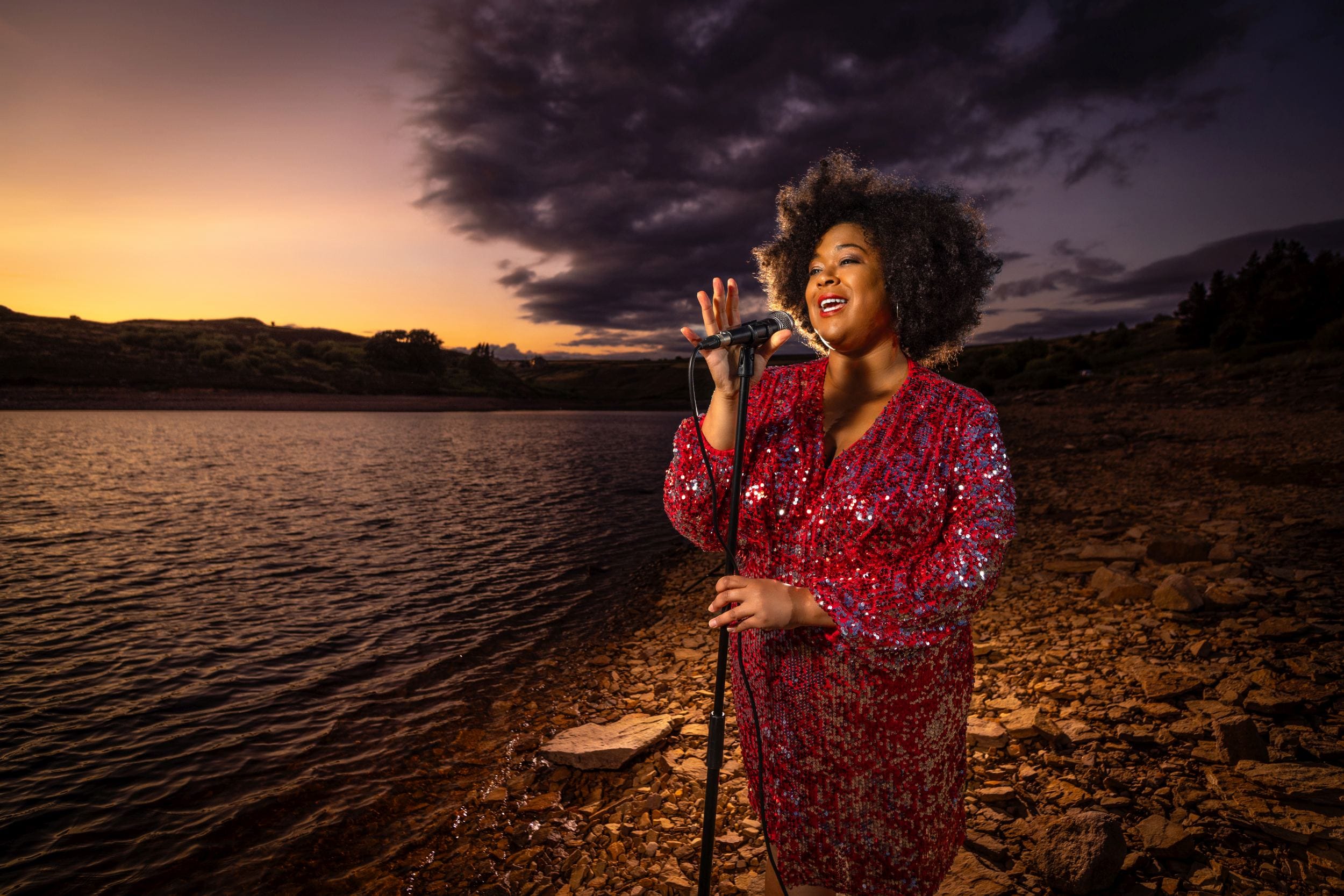 Woman in a red sequin dress sings passionately into a microphone by a lake at sunset with dramatic clouds in the background.