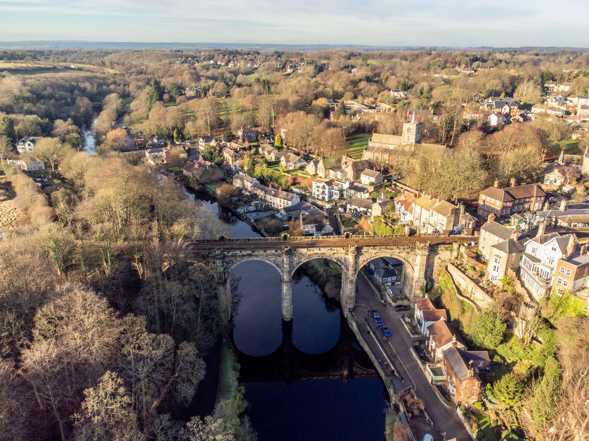 Aerial view of a railway viaduct crossing a river, surrounded by a village with houses, trees, and open fields.