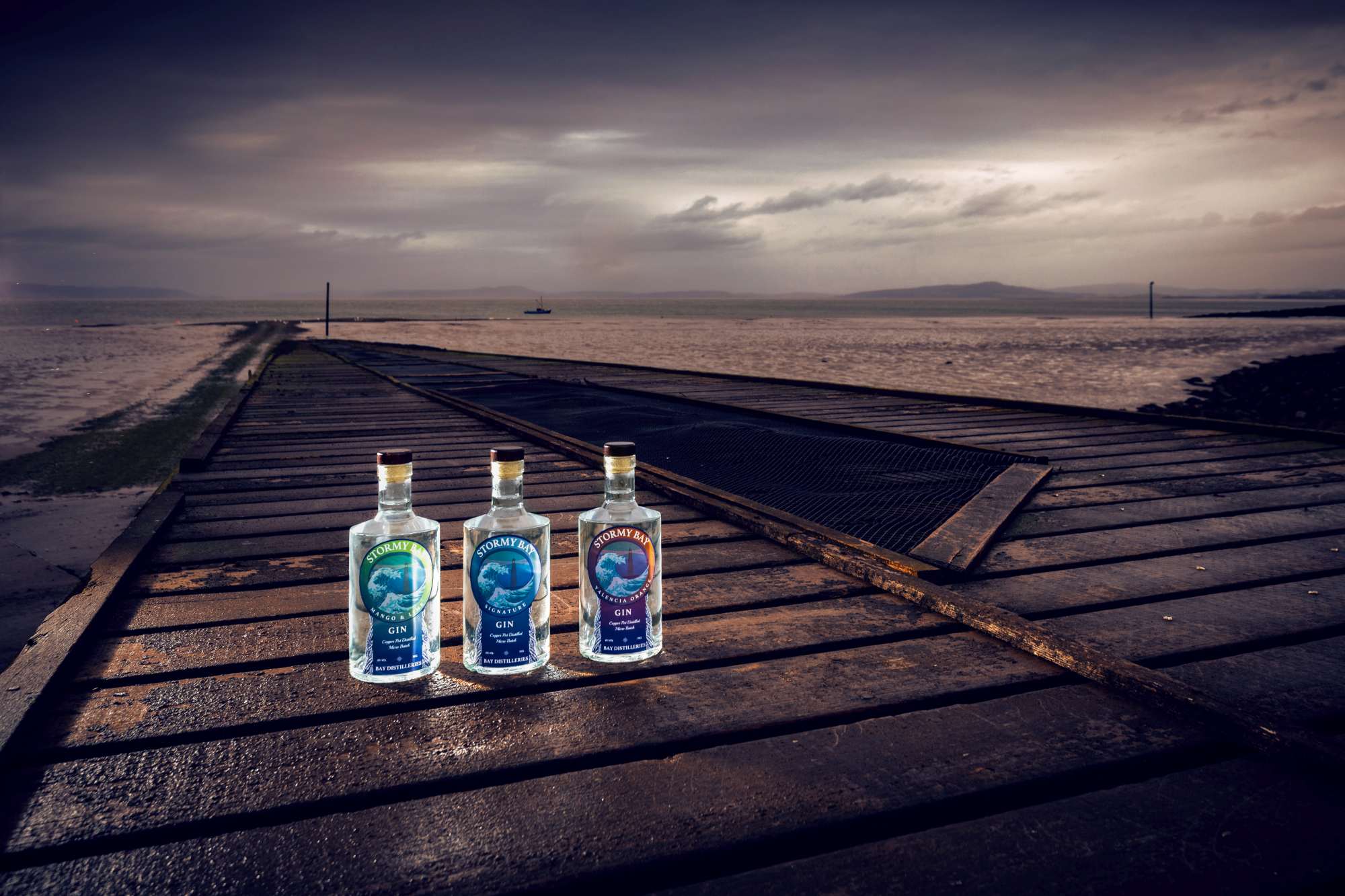 Three bottles of Stormy Bay gin on a weathered wooden pier with a stormy seascape in the background.