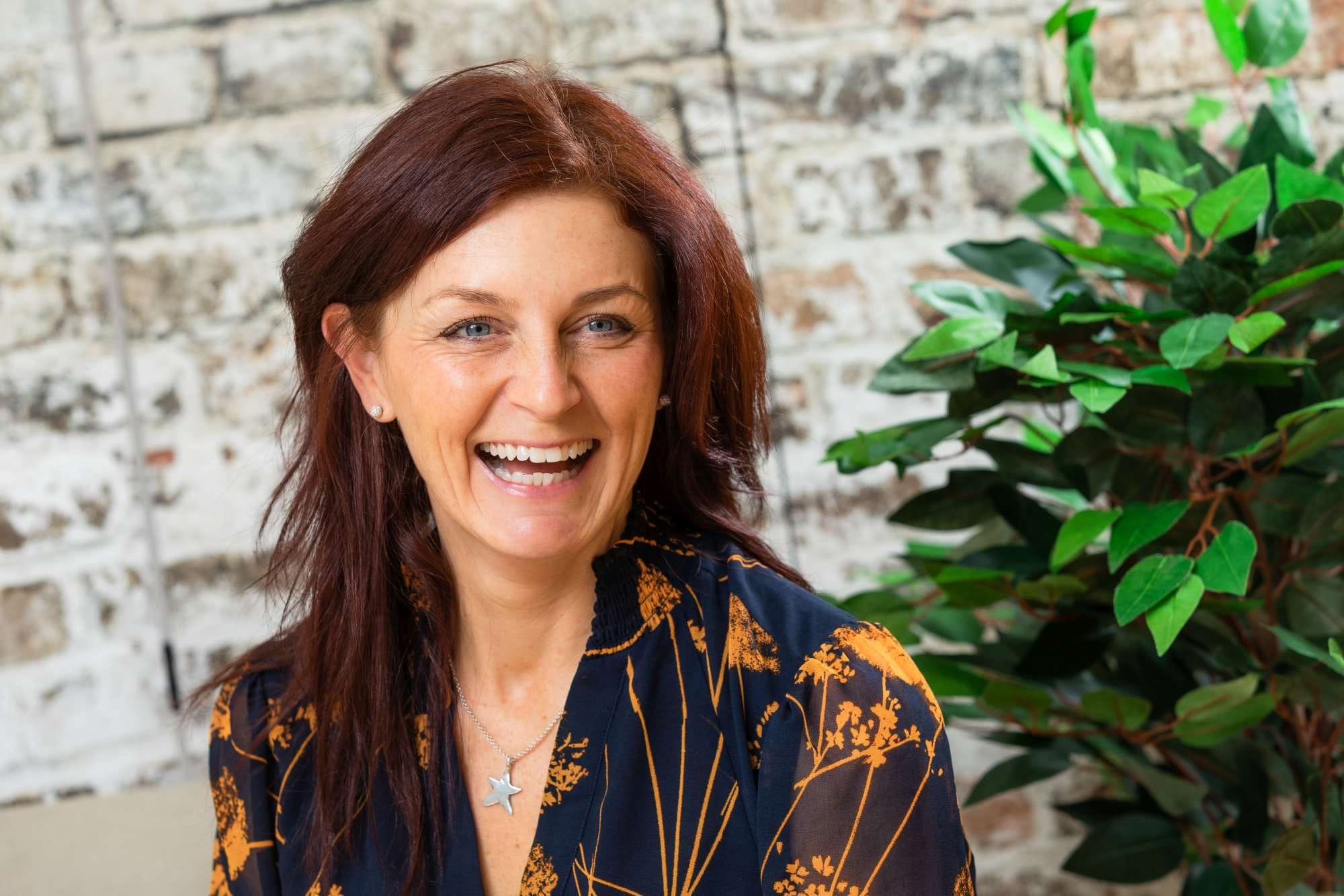 Woman with auburn hair laughing, wearing an orange and black floral blouse, sitting indoors with a brick wall and plant background.