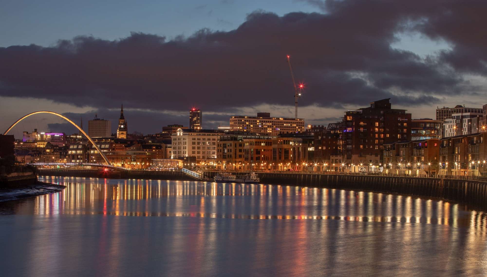 City skyline at dusk with illuminated buildings reflecting on a river, featuring a prominent bridge and a crane in the background.