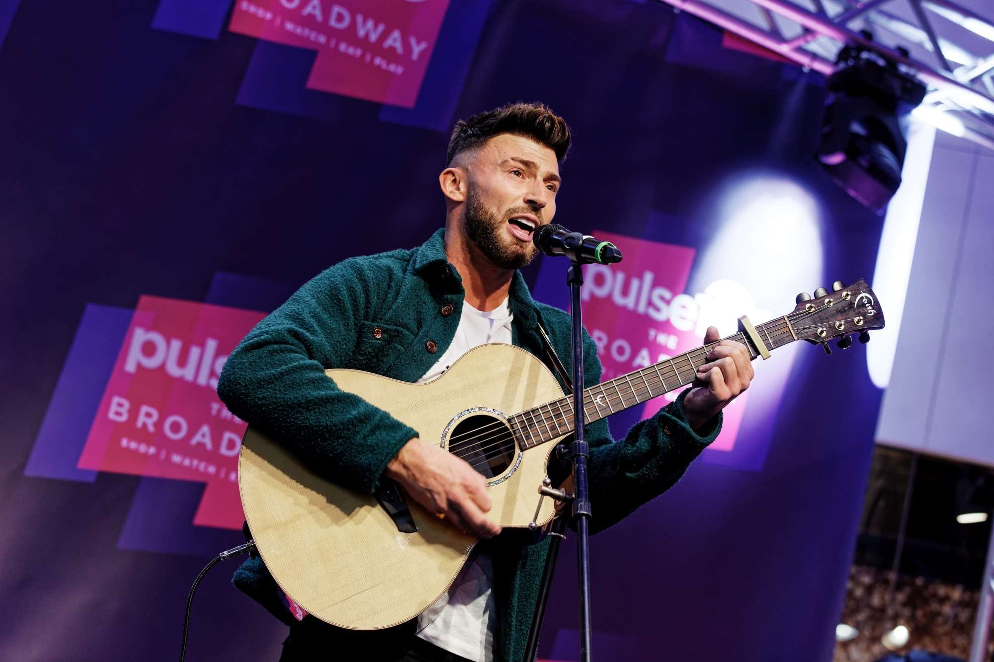 Musician performing on stage with an acoustic guitar, wearing a green jacket, with a purple banner background that reads "Pulse" and "Broadway.