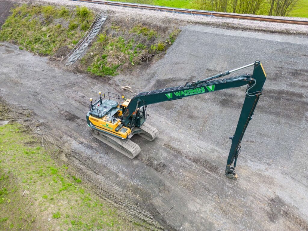 A long-reach excavator works on a gravel slope near a railway track, with a metal staircase visible on the embankment.