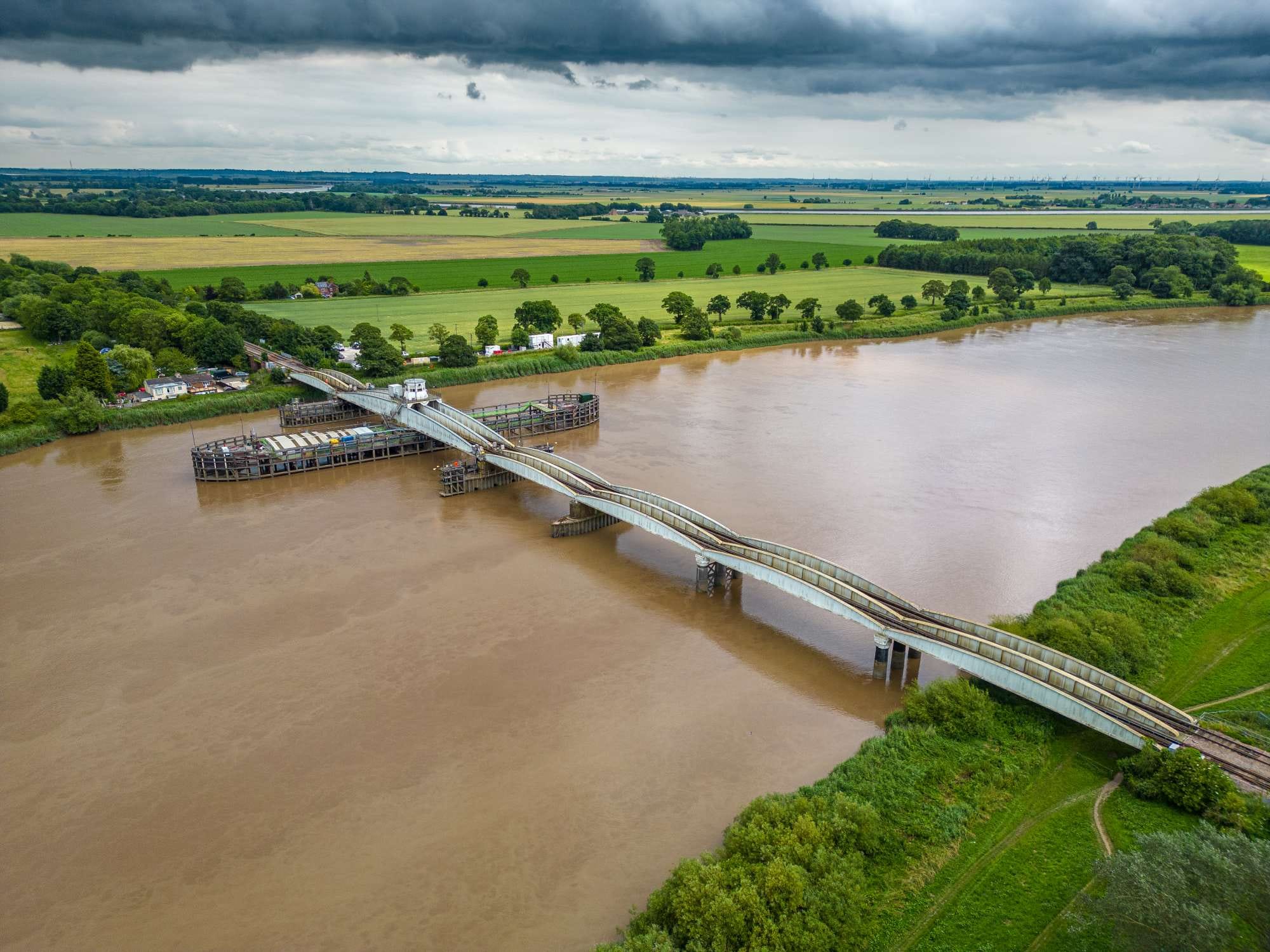 Aerial view of a partially constructed bridge over a wide river with surrounding green fields and overcast sky.