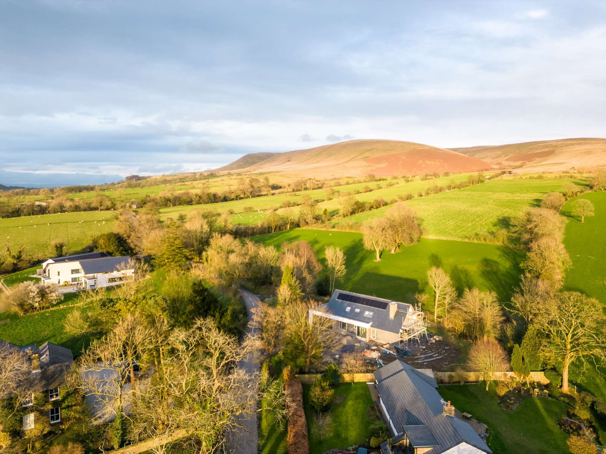 Aerial view of a rural landscape with green fields, scattered trees, and several houses, under a partly cloudy sky.