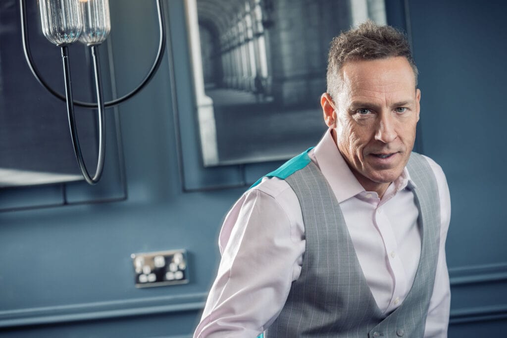 Man in a light gray vest and light pink shirt stands in a blue room with modern decor and framed pictures.