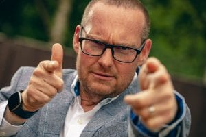 A man wearing eyeglasses and a blazer points at the camera with both hands, wearing a focused expression.