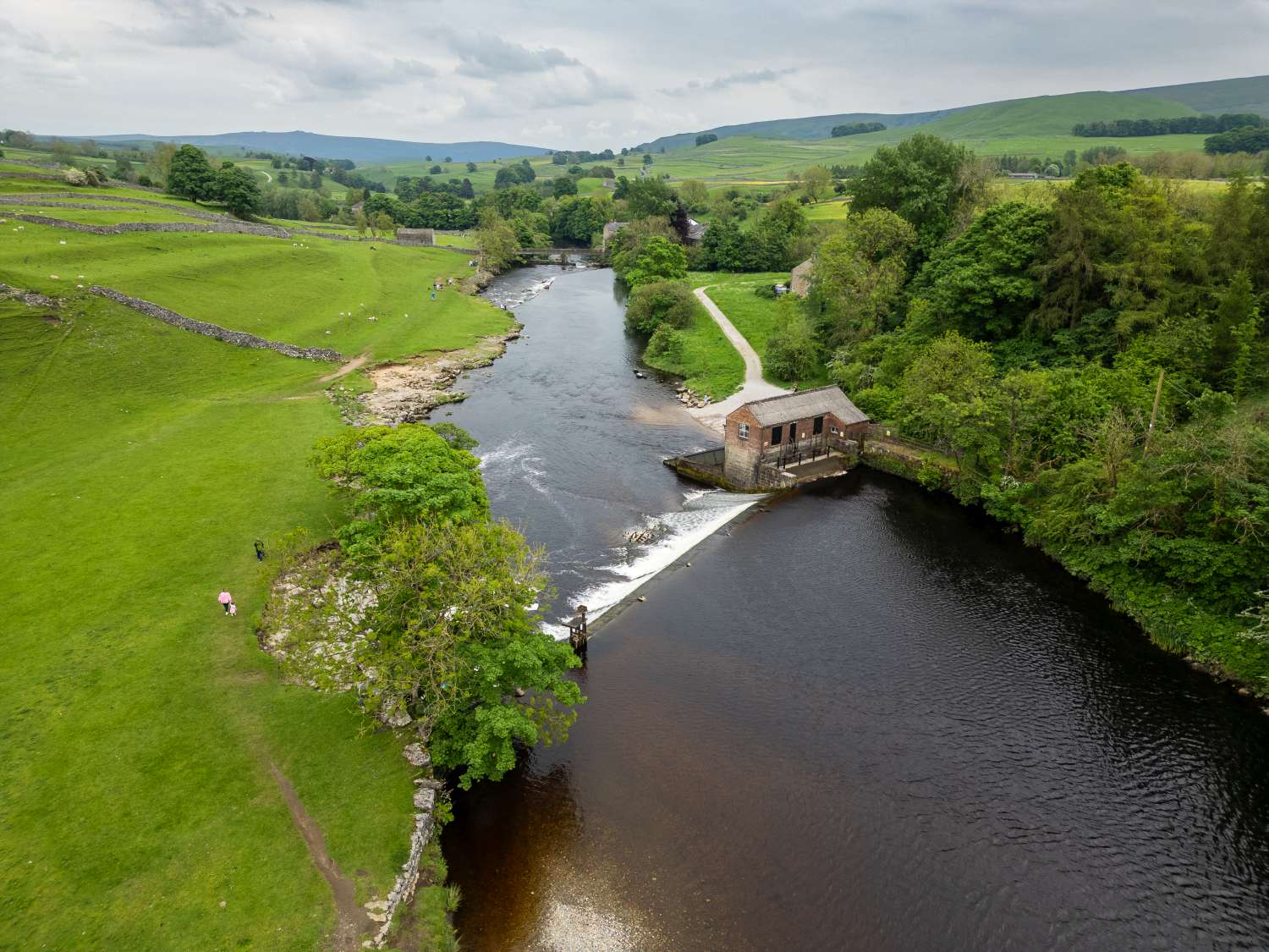 Aerial view of a river with a small dam and a brick building, surrounded by green fields, trees, and rolling hills.