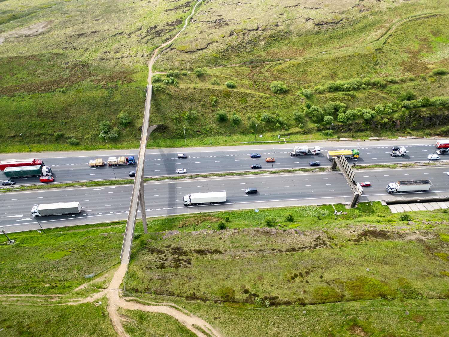 Aerial view of a highway with multiple lanes, vehicles, and a pedestrian footbridge crossing over it surrounded by green fields.
