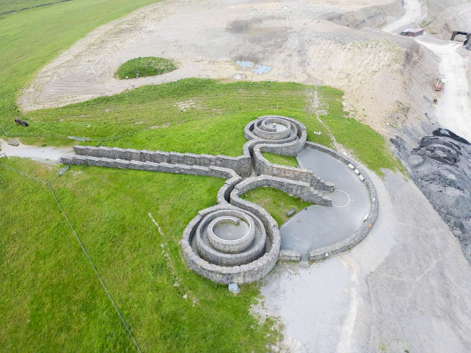 Aerial view of sculpted stone labyrinth on green landscape, featuring intricate spiral pathways and surrounding open field.