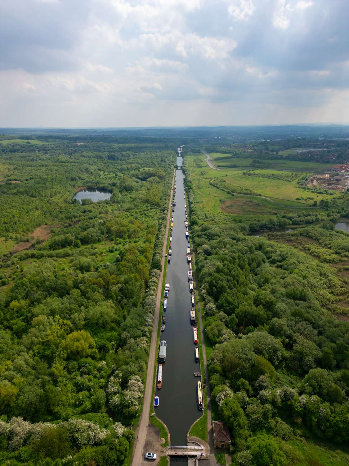 Aerial view of a long canal with numerous boats surrounded by lush greenery and fields, under a partially cloudy sky.