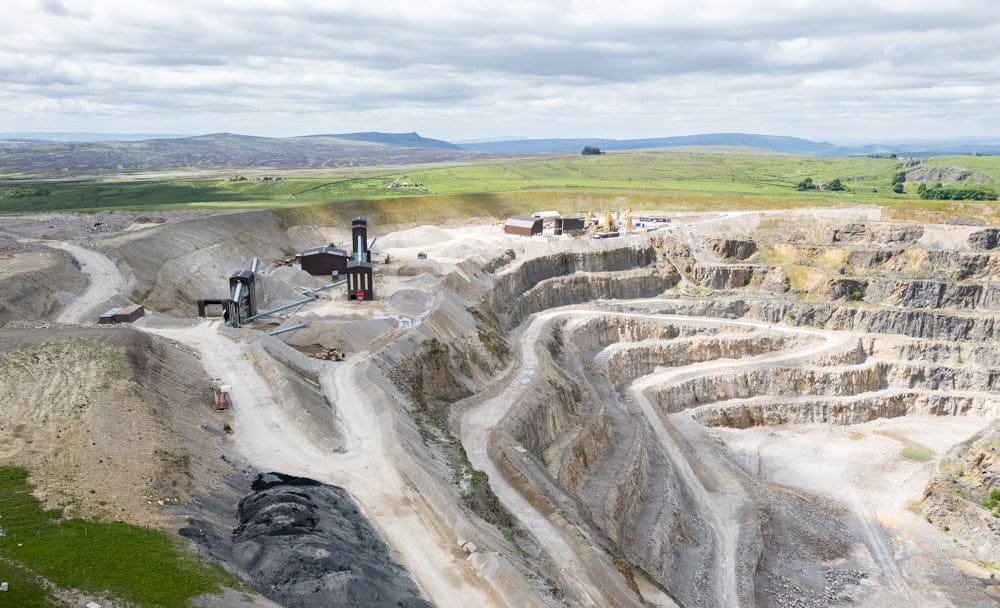 A large limestone quarry with terraced layers and processing buildings under a cloudy sky, set in a rural landscape.