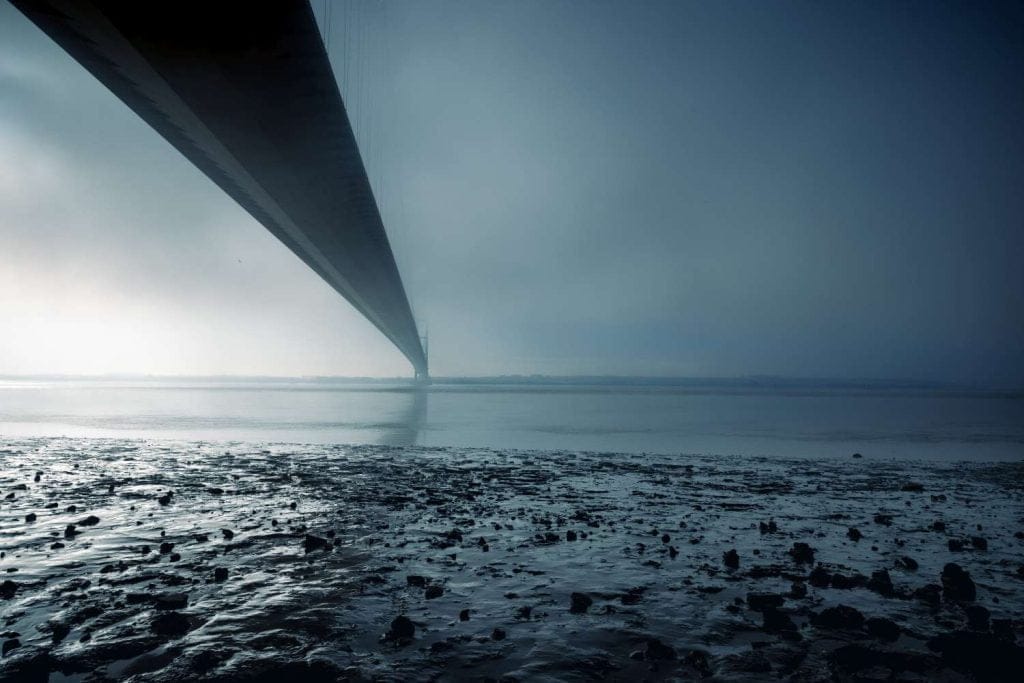 Long bridge extends over calm waters, shrouded in mist, with a rocky shoreline in the foreground.