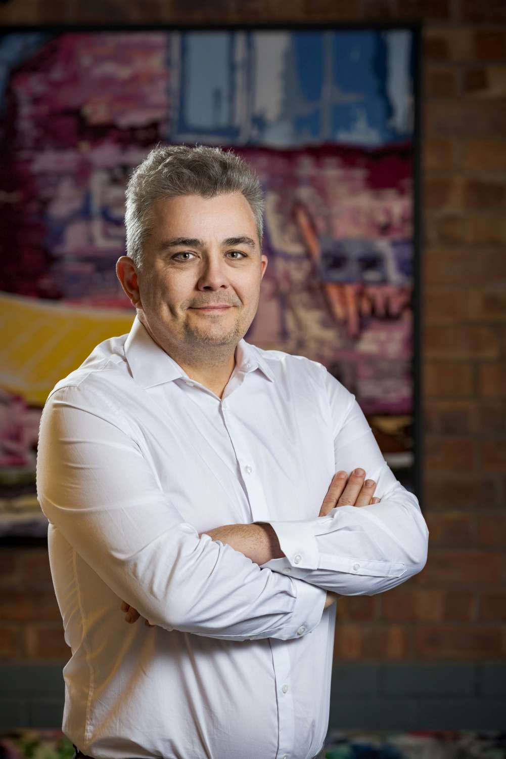 Man in white shirt with arms crossed, smiling in front of colorful abstract artwork and a brick wall background.