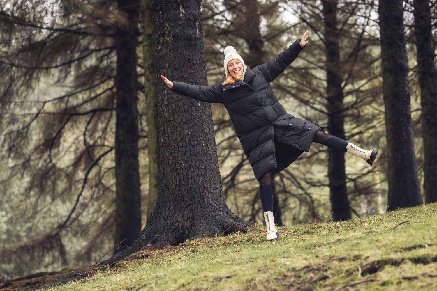 Person in a black coat and white hat playfully balancing on one leg next to a tree in a forested area.