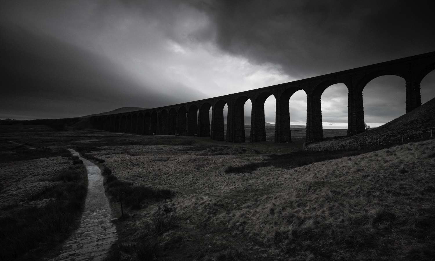 Stone viaduct with multiple arches stretches across a grassy landscape under a dramatic, cloudy sky. A winding path leads towards it.
