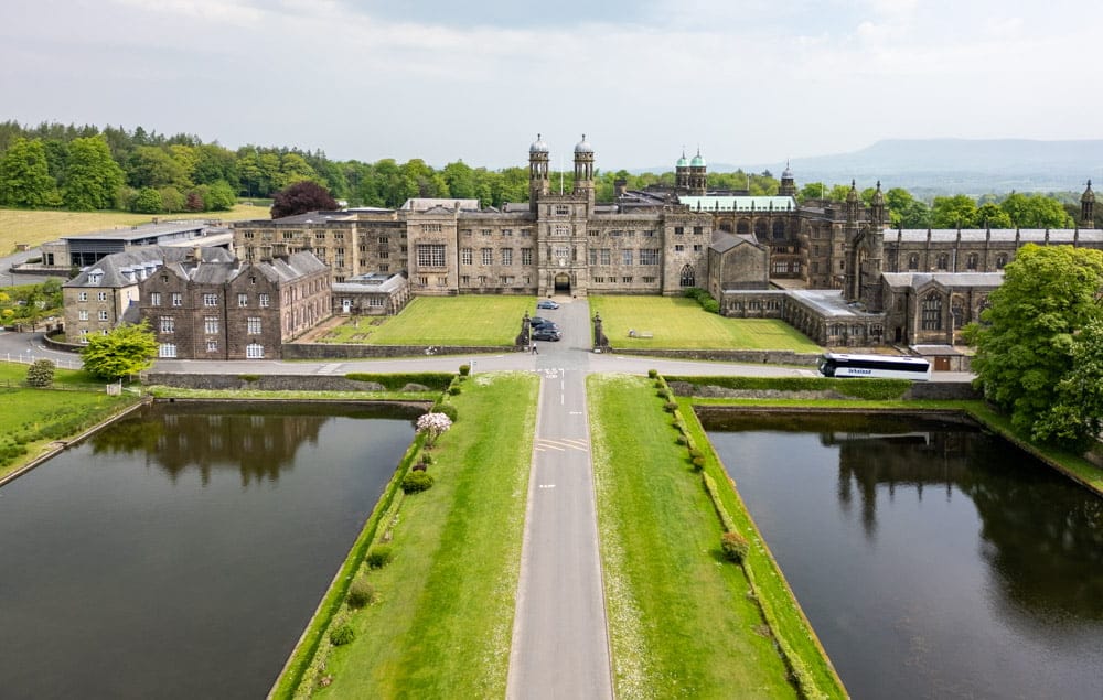 Aerial view of a historic stone building with towers, surrounded by lush greenery and reflective ponds.