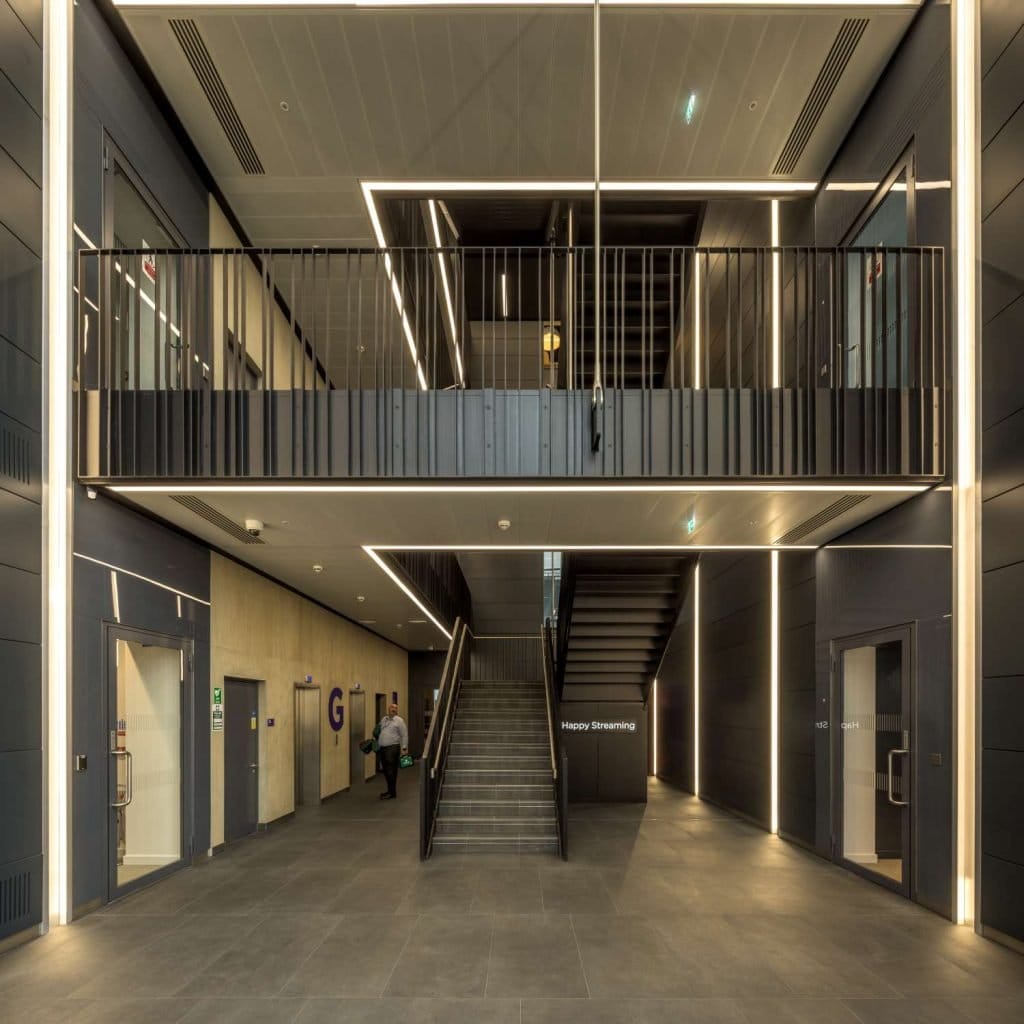 Modern lobby with dark tones, central staircase, upper-level balcony, and person walking towards door marked "G" on the left.