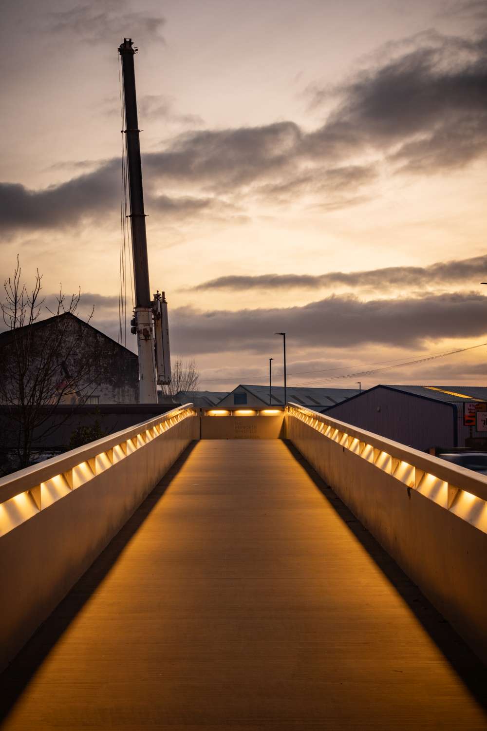 Illuminated pedestrian bridge at sunset with buildings and a crane in the background. Sky is partly cloudy.