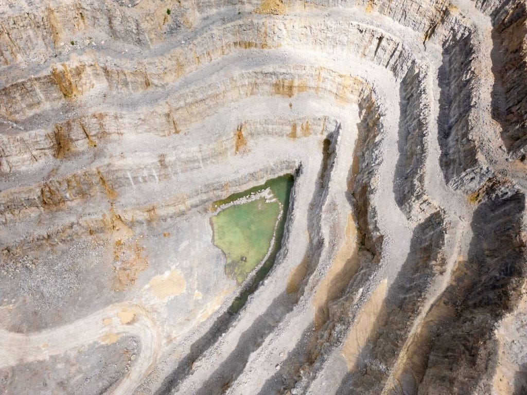 Aerial view of a stepped quarry with a small greenish water pool at the bottom, surrounded by layered rock formations.