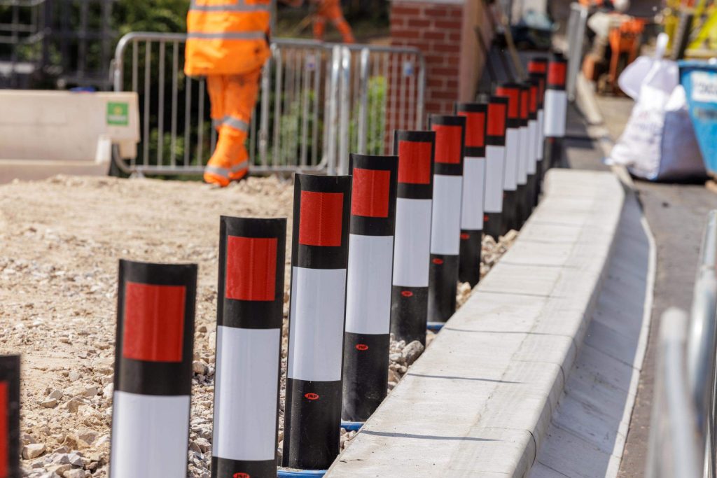 Red and white safety bollards lining a construction site walkway, with a worker in hi-vis in the background