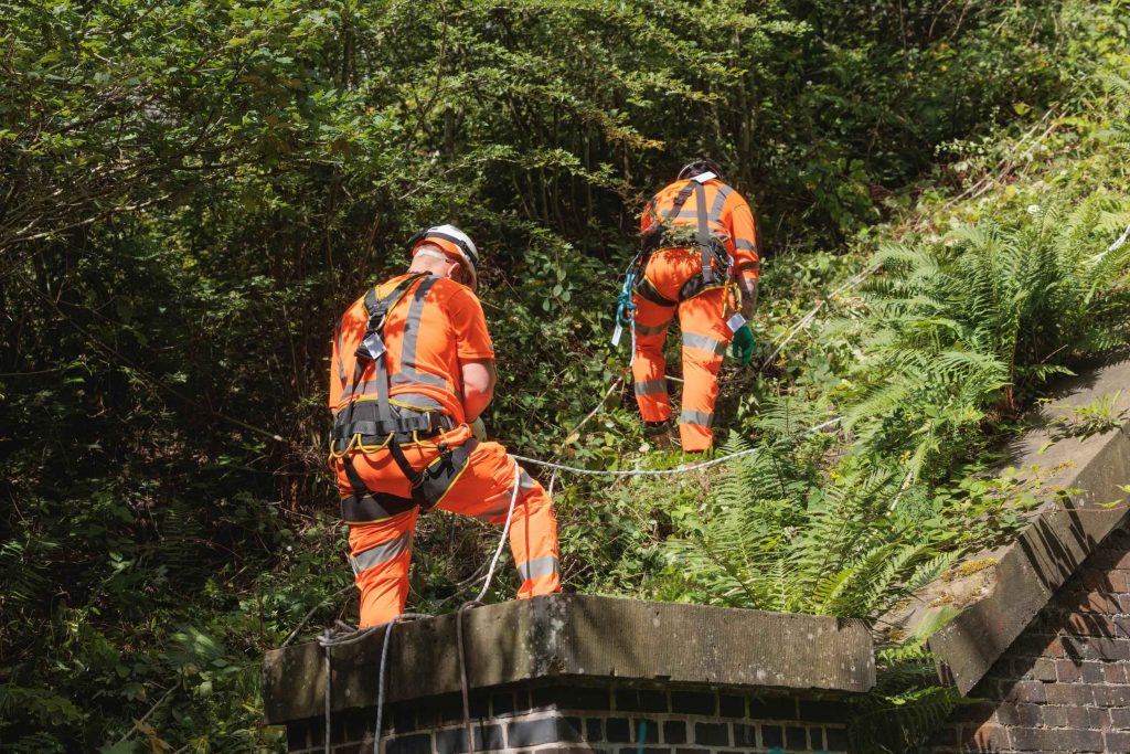 Two site workers in harnesses and hi-vis working on a bridge structure set into a wooded embankment