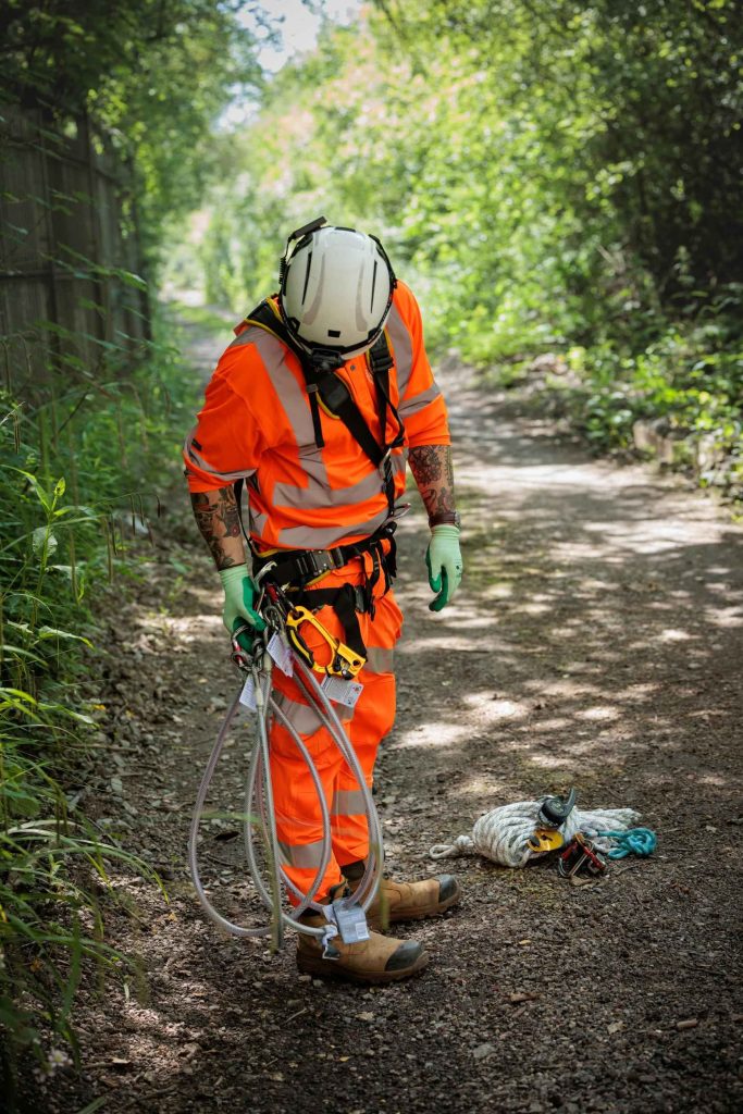Site worker in hi-vis and harness walking along a rural access path carrying equipment