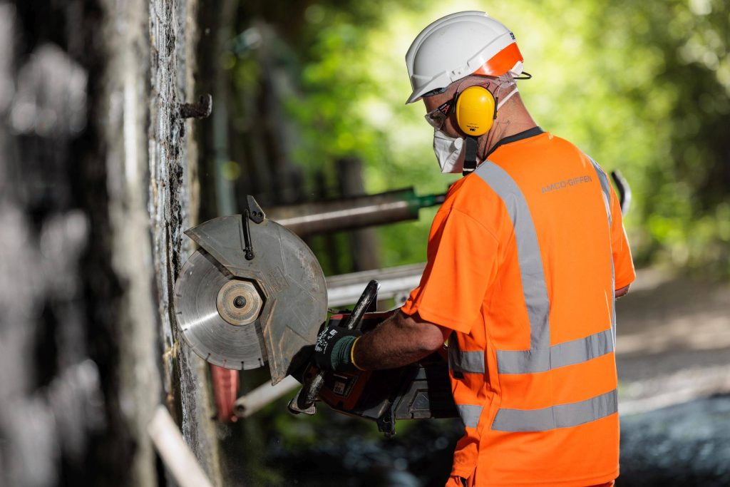 Close-up of a worker in hi-vis using a power cutter on a concrete pipe section, dust visible in the air