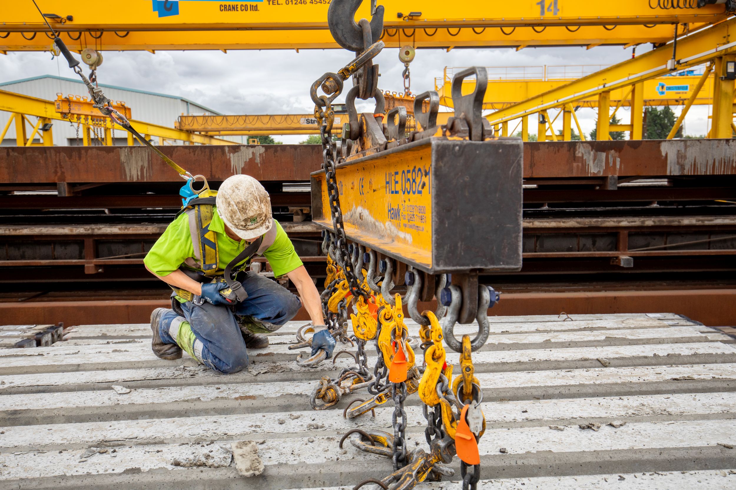 Site worker clipped into a harness attaching lifting chains to a steel beam under a gantry crane