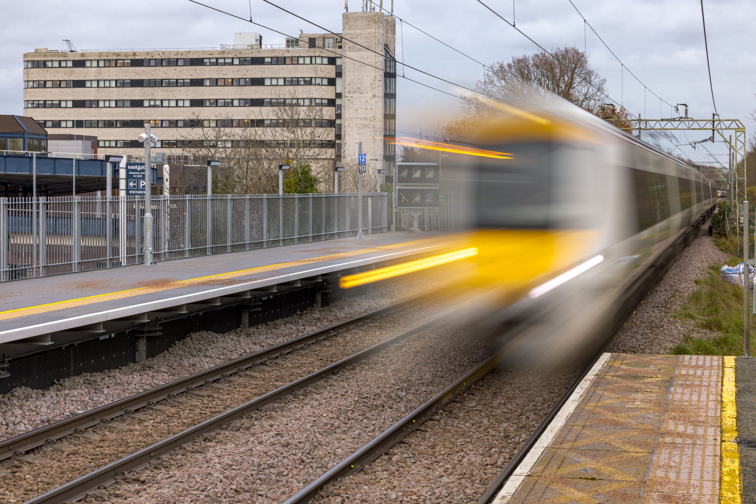 A train departs a railway station platform in motion blur, with an office building in the background