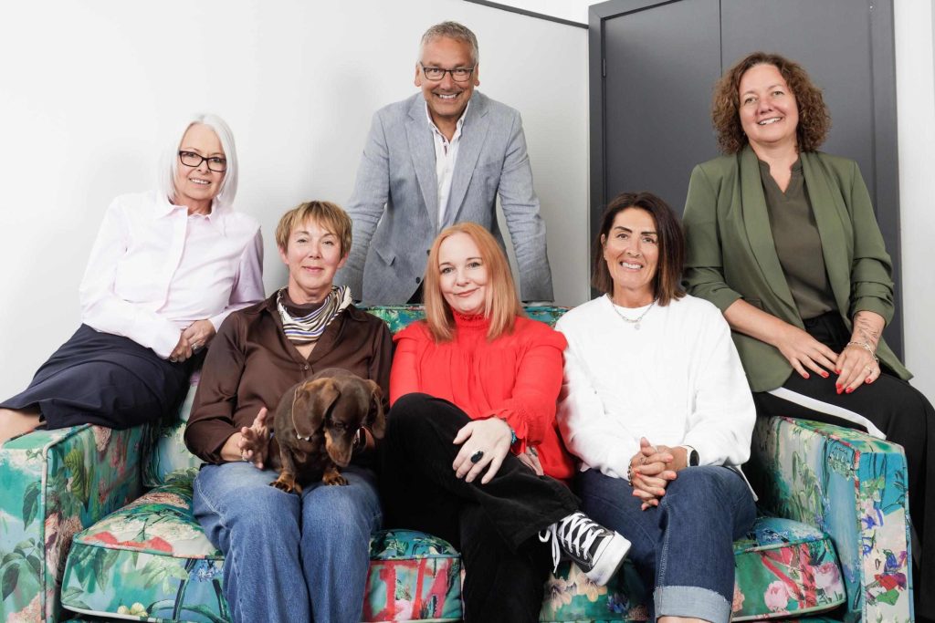 Group team portrait of six colleagues relaxed on a patterned sofa, with a dachshund dog
