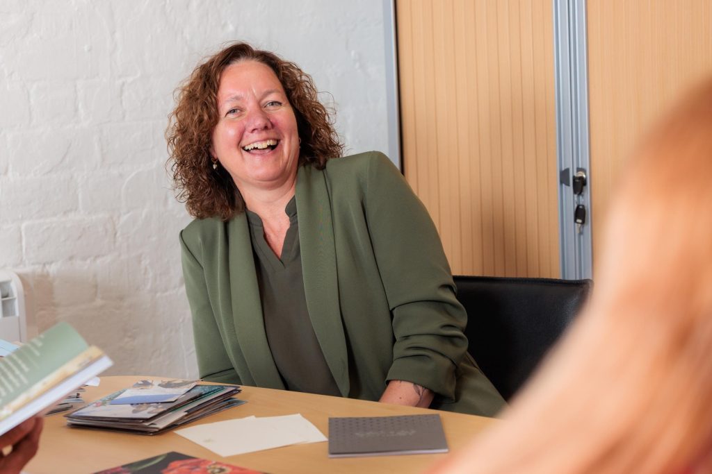 Woman in a green blazer laughing during a meeting, seated at a table with documents