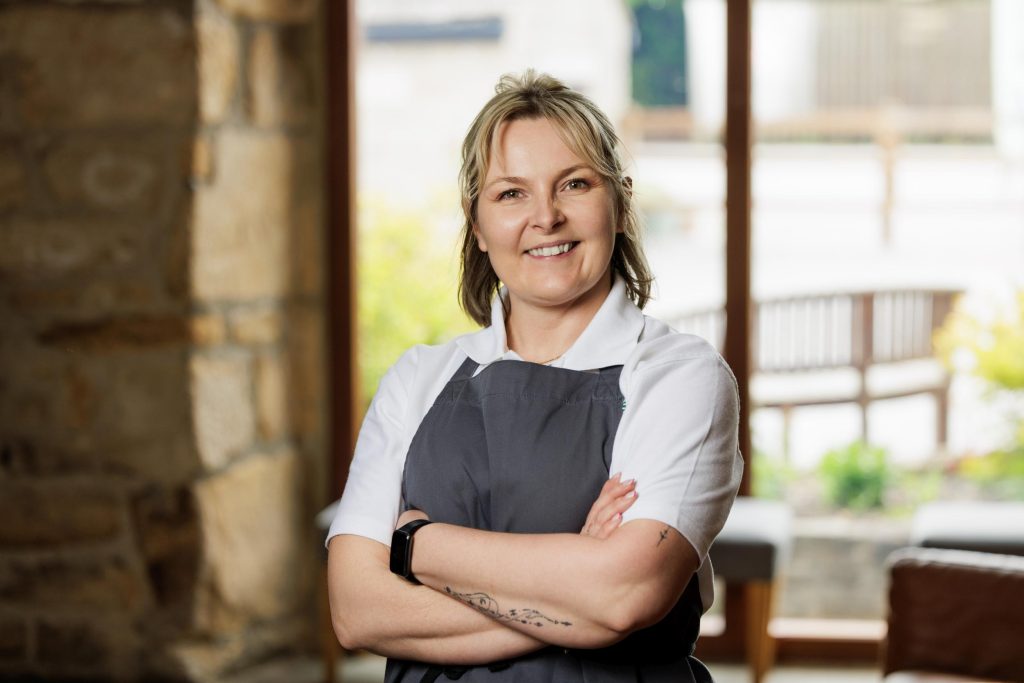 Woman in an apron standing arms crossed by a stone wall, smiling