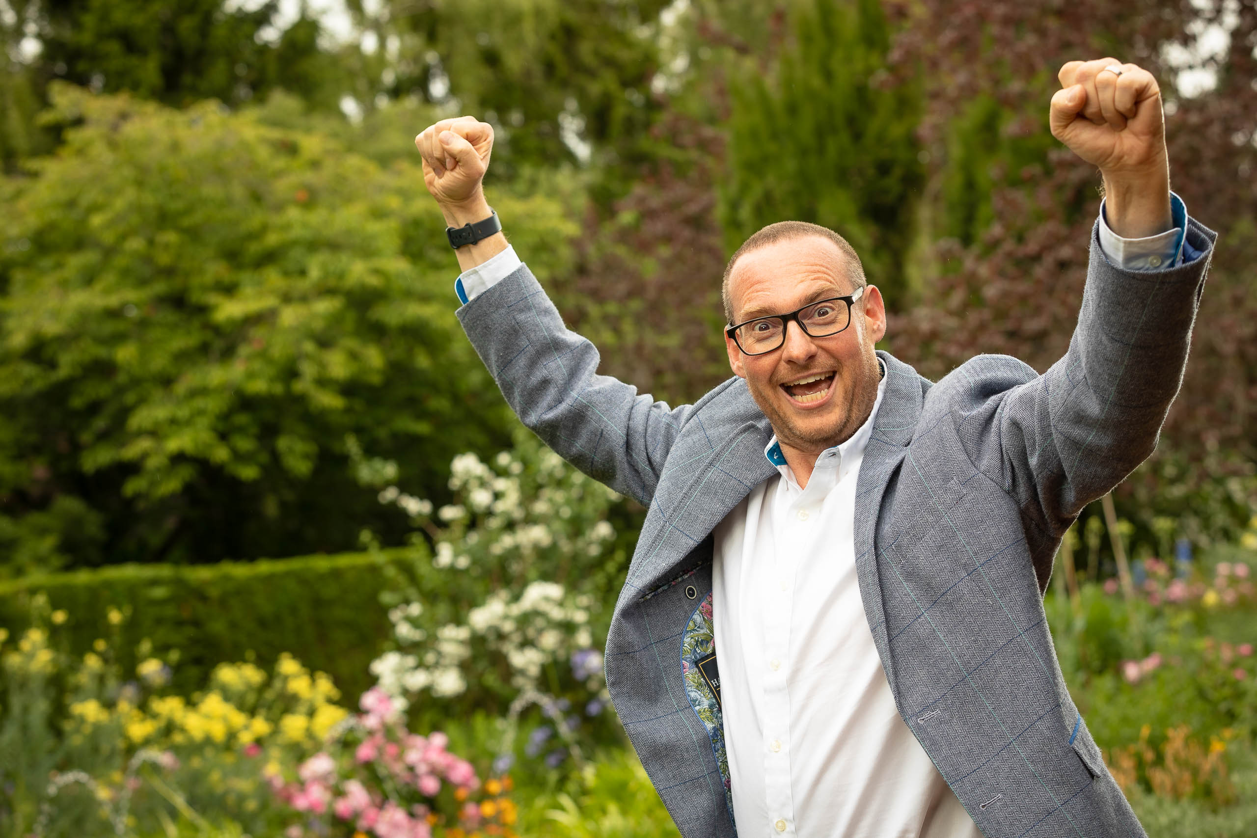 Man wearing glasses and a blue jacket raises his fists and smiles excitedly in a colorful garden with various flowers and greenery.