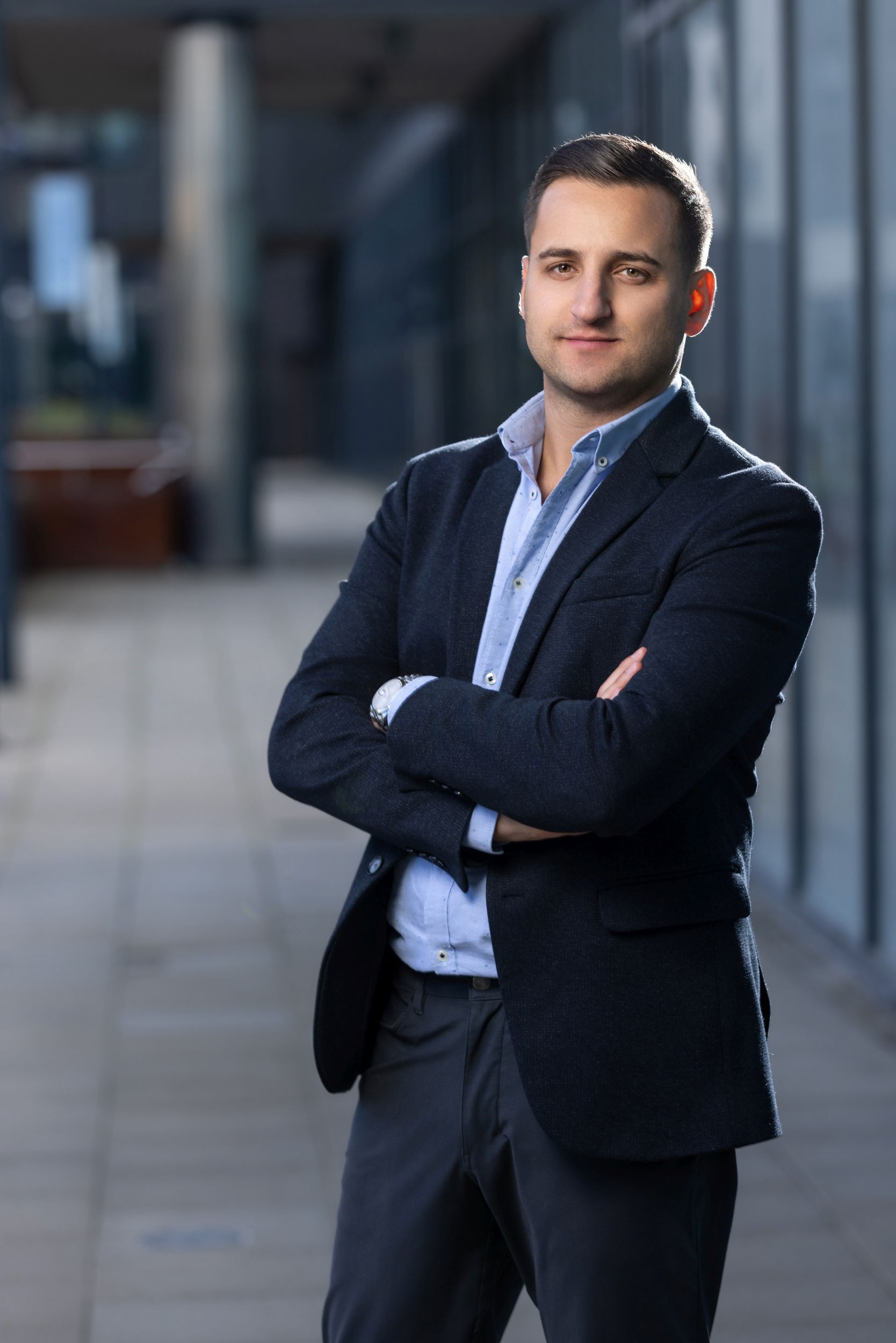 Confident man in a navy blazer, arms crossed, in an urban office corridor