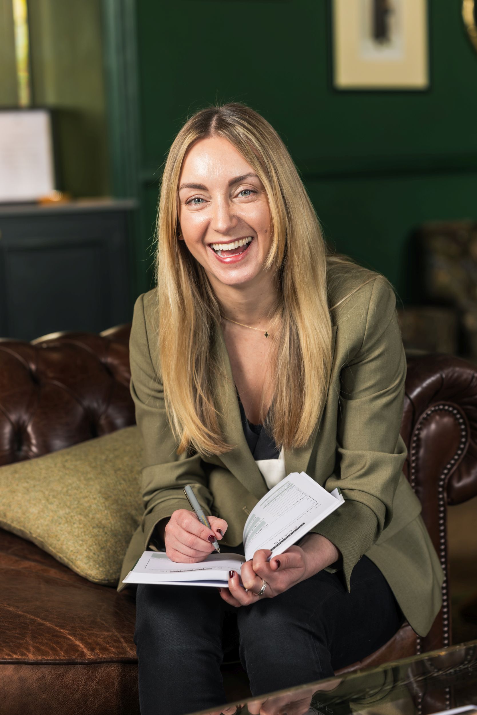 Woman laughing while making notes in a notebook on a leather sofa, styled office setting